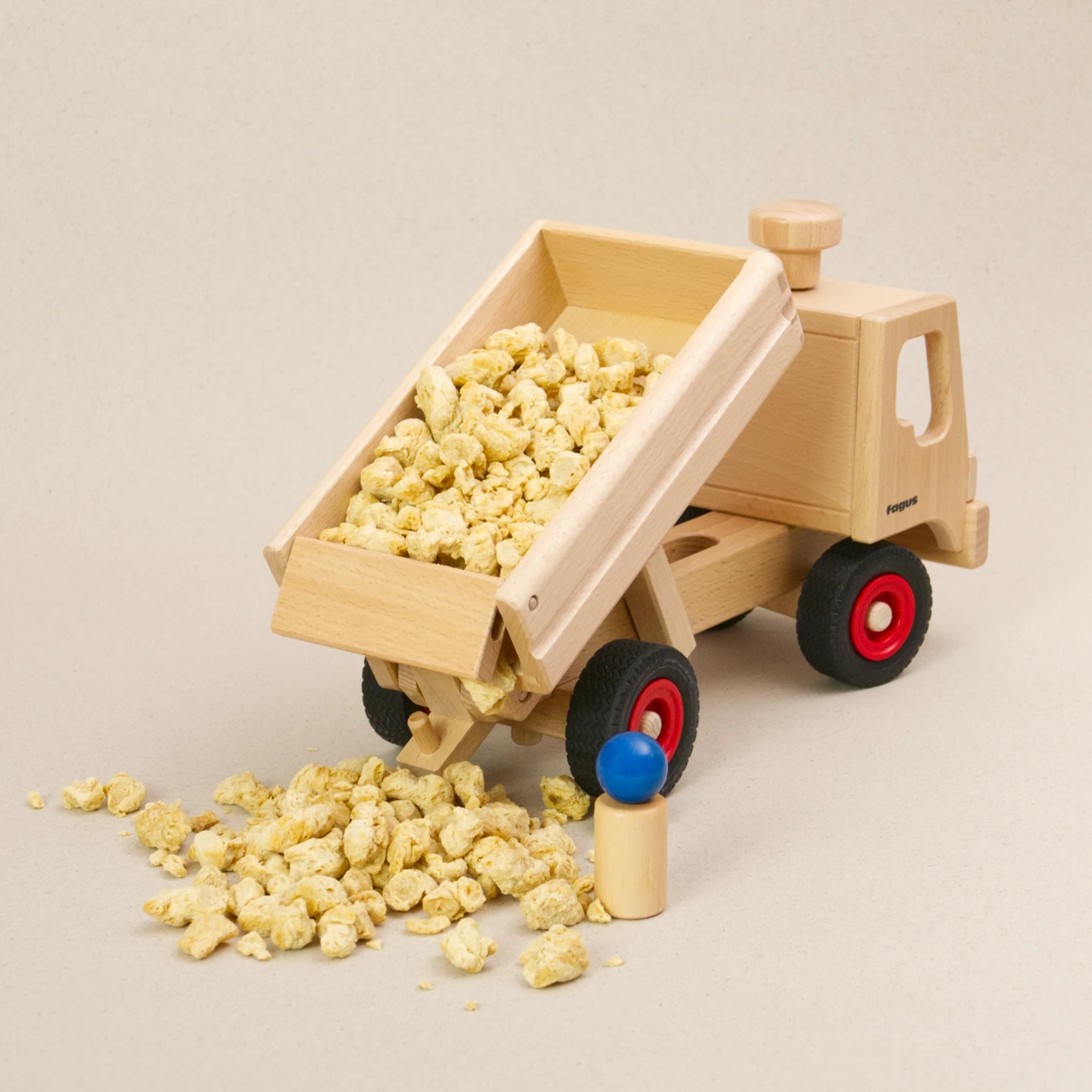 Holzfahrzeug LKW Kipper von fagus aus naturlackiertem Buchenholz mit Vollgummireifen. Ansicht von hinten rechts, die mit Schüttgut gefüllte Ladefläche ist nach oben gekippt und die Ladeklappe ist geöffnet, sodass es raus geschüttet wird. Eine Holzfigur beobachtet den Vorgang.