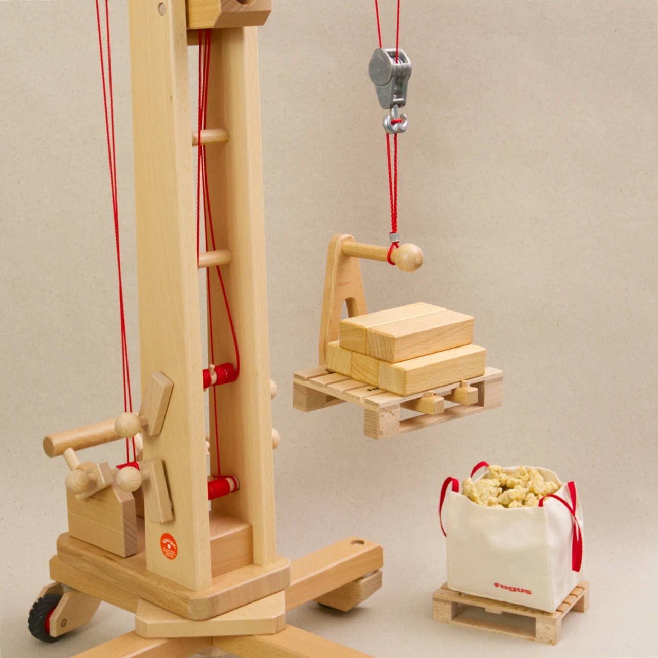 Ladegabel von fagus aus naturlackiertem Buchenholz mit roter Verbindungsschnur am Haken vom fagus Hochkran angebracht. Die Ladegabel schwebt nah über dem Boden und ist mit einer Europalette mit Holzblöcken beladen. Eine Europalette mit gefülltem Big Bag steht daneben.