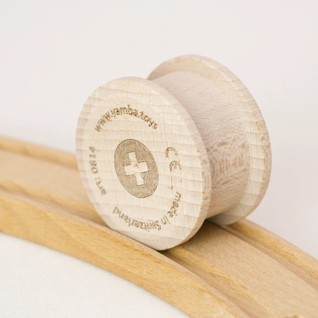 Die Holzwalze Rolli von der Marke Yamba ist in einer Nahaufnahme auf einer Spielzeugeisenbahnschiene aus Holz zu sehen. Der Rolli hat die Form eines Zylinders, der an den Enden jeweils etwas Überstand hat, um in der Schiene laufen zu können. Der Rolli ist aus naturbelassener Schweizer Buche und aus einem Stück gefertigt und verzichtet auf Verwendung von Leim und Lack.