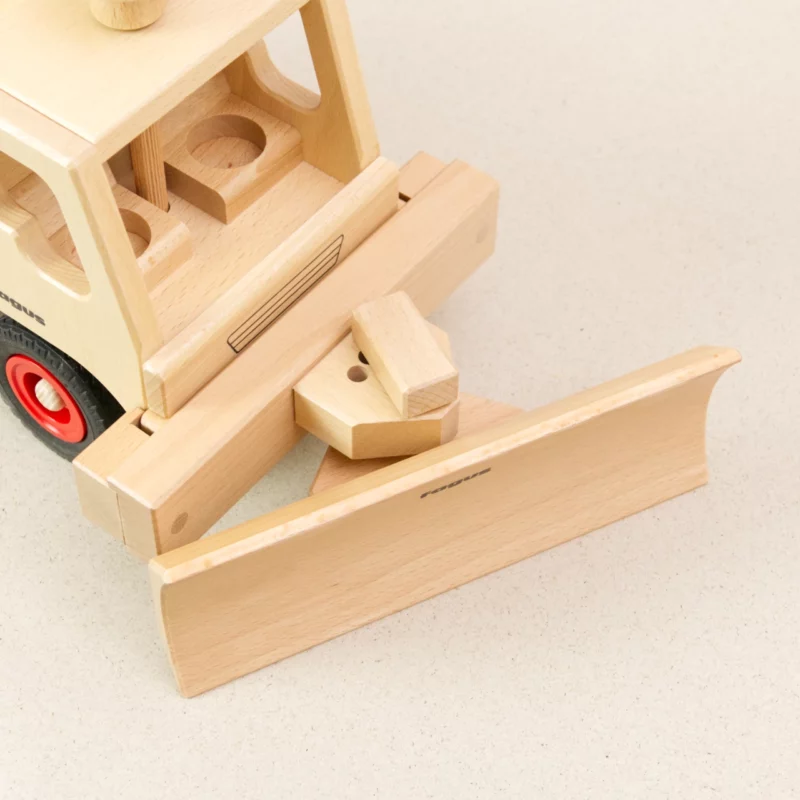 Schneepflug von fagus aus naturlackiertem Buchenholz, mit schwarzen Vollgummireifen und roten Radkappen. Die Fahrerkabine ist leer und der Schneeschieber ist an das Fahrzeug gekoppelt und nach links ausgerichtet.