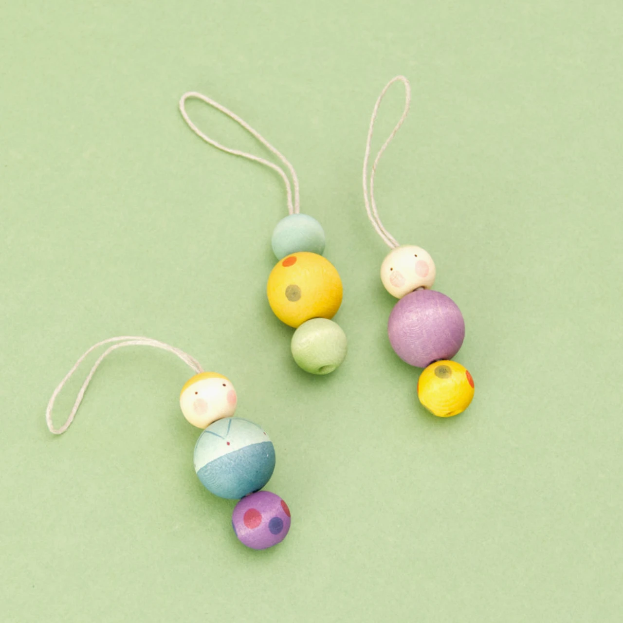 Die drei Figuren der Serie Snowy Buddy von den Grapat Ornaments liegen auf einem grünen Untergrund. Alle Weihnachtsbaumanhänger bestehen aus drei Holzkugeln. Der mittig platzierte Anhänger ist bunt gemustert, während die beiden anderen Figuren ein Gesicht haben. Die Anhänger sind in Violett, Gelb, Blau und Grün gestaltet, mit weiteren farblichen Akzenten.