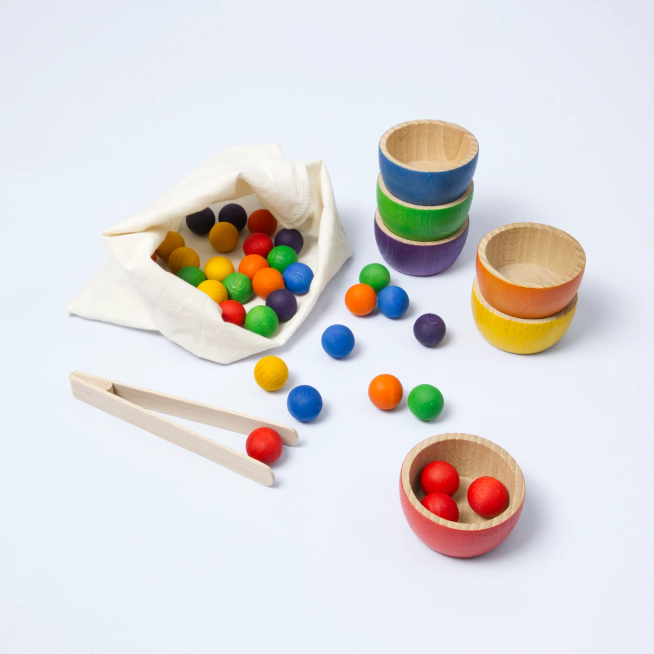 Säckchen mit verschiedenen kleinen Holzkugeln in 6 verschiedenen Farben, daneben gestapelte Holzbecher in auch je 6 Farben und eine Zange um die kleinen Kugeln in die Becher nach Farbe zu sortieren