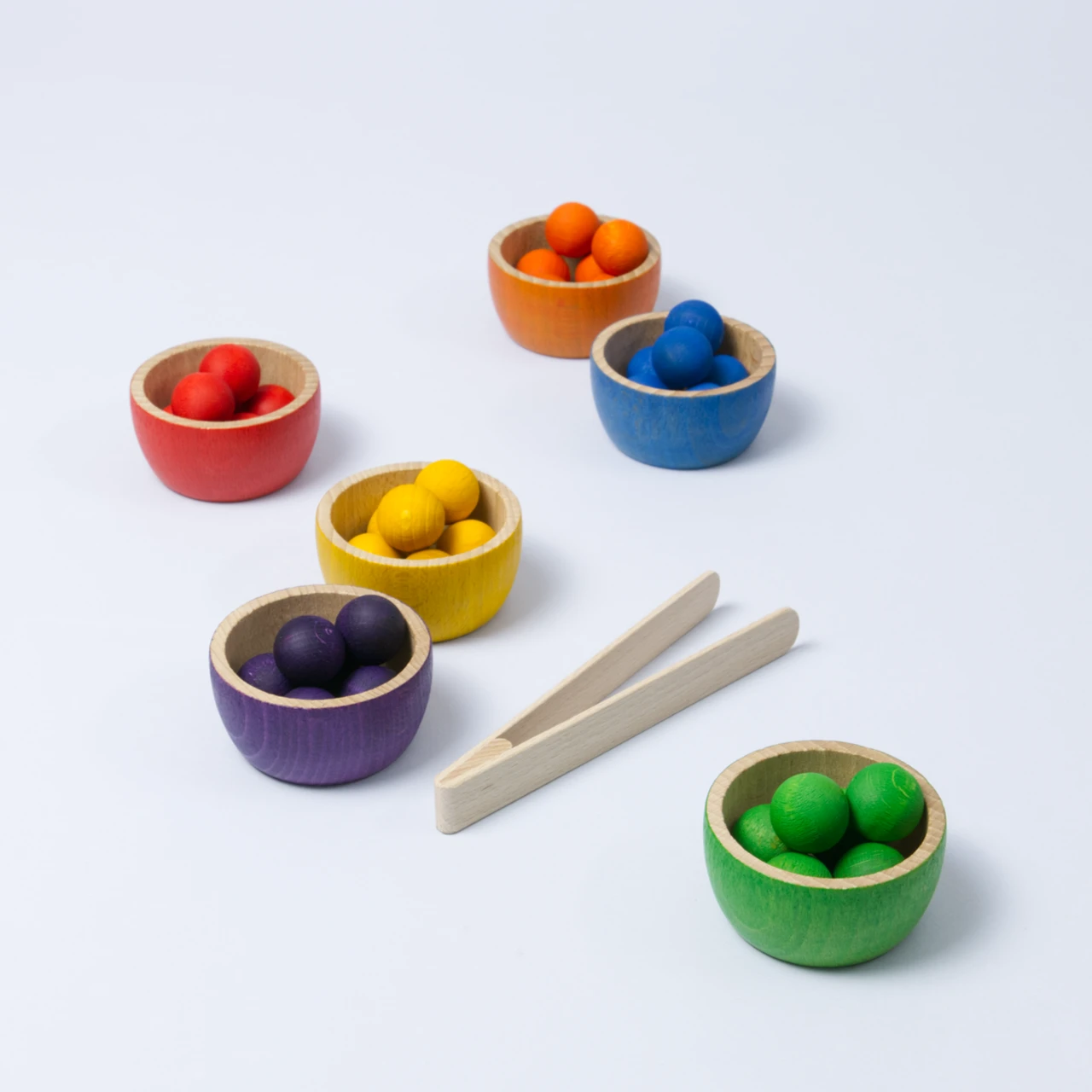 6 kleine Schüsseln in Regenbogenfarben, gefüllt mit 6 farbig passenden Kugeln, sowie eine Zange. Alles aus Holz.