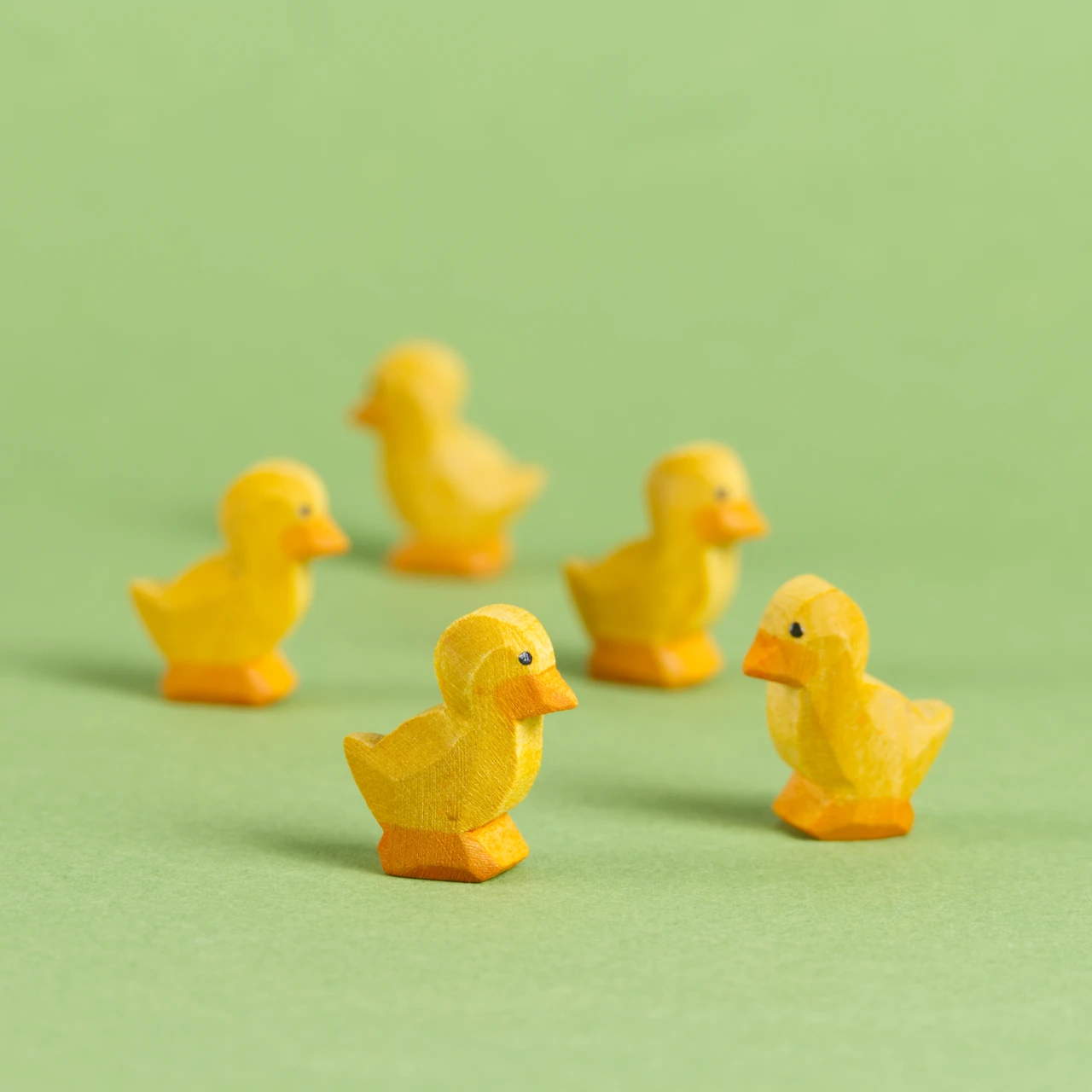 Komposition aus fünf Entenküken von Ostheimer aus Naturholz gefertigt. Alle haben gelbes Fell, einen orangenen Schnabel sowie Paddeln. Das Auge ist durch einen schwarzen aufgemalten Punkt gekennzeichnet. Zwei im Vordergrund schauen sich gegenseitig an, zwei in der Bildmitte schauen nach rechts und das im Hintergrund nach links.