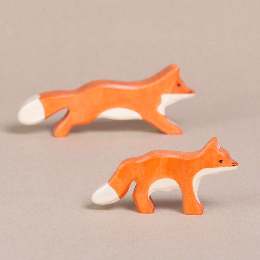 Komposition aus zwei nach rechts schauenden Füchsen von Holztiger, beide haben orangenes Fell und eine weiße Schwanzspitze und weißes Bauchfell. Die Ohren stehen gerade nach oben. Eines ist klein das andere laufend.