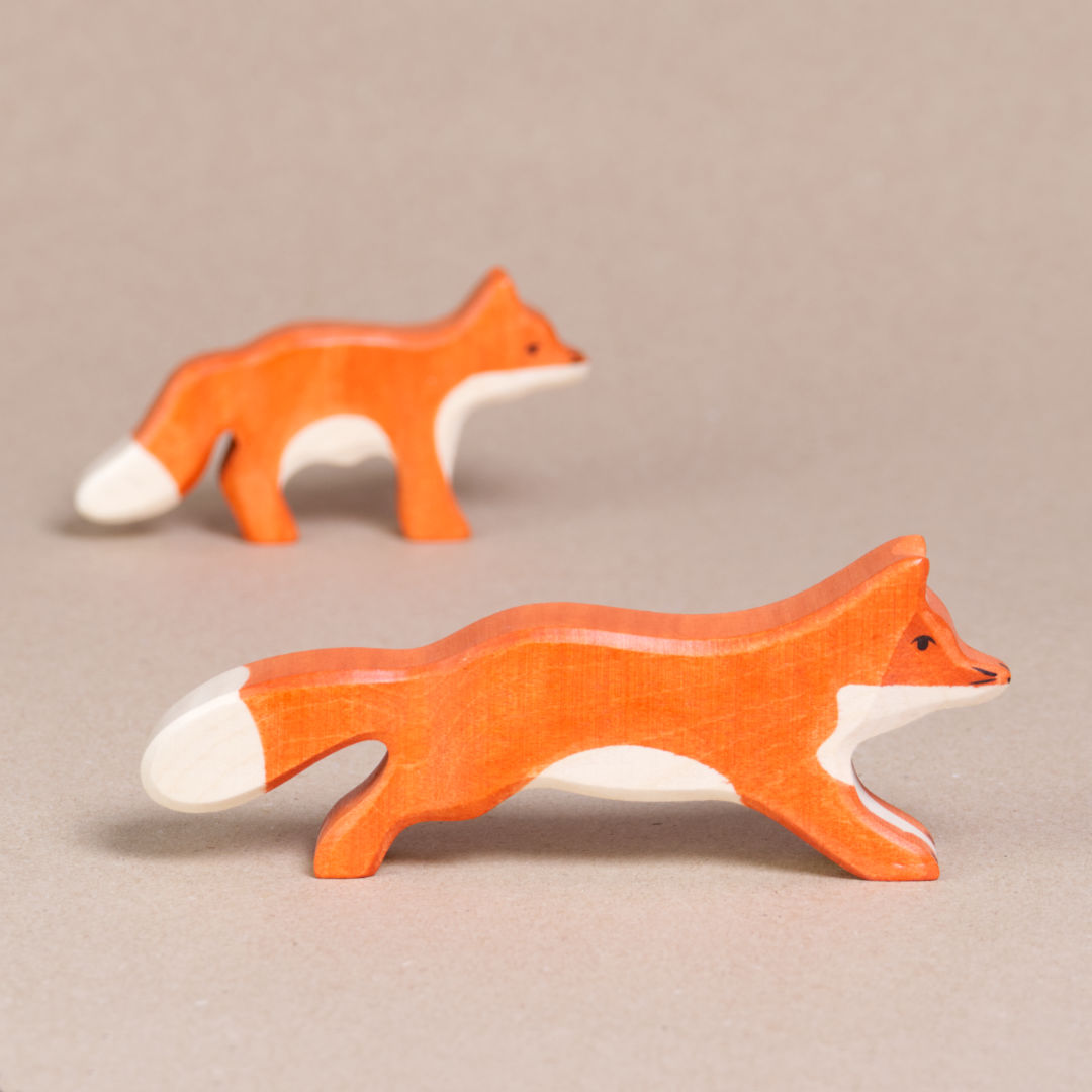 Ein nach rechts schauender, grosser, laufender Fuchs von Holztiger, ist orange und hat eine weiße Schwanzspitze und weißes Bauchfell. Die Ohren stehen gerade nach oben. Im Hintergrund ist der kleine Fuchs zu sehen.