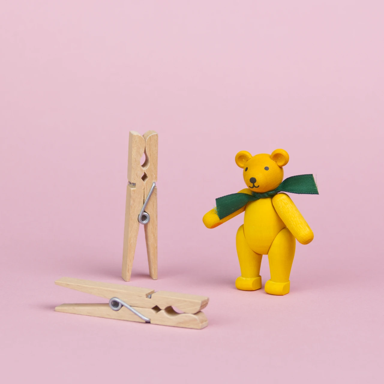 ein kleiner Teddybär aus gelb bemaltem Holz mit einer grüner Schleife um den Hals steht neben 2 Wäscheklammern, die beiden Objekte sind annähernd gleich groß, seine Augen und Nase sind schwarz und sein Mund lächelt