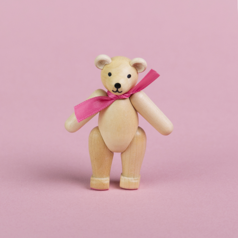 ein kleiner Teddybär aus hellem Holz mit rosa Schleife um den Hals, Beine und Arme sind beweglich und er ist in einer stehenden Position zu sehen, seine Augen und Nase sind schwarz und sein Mund lächelt