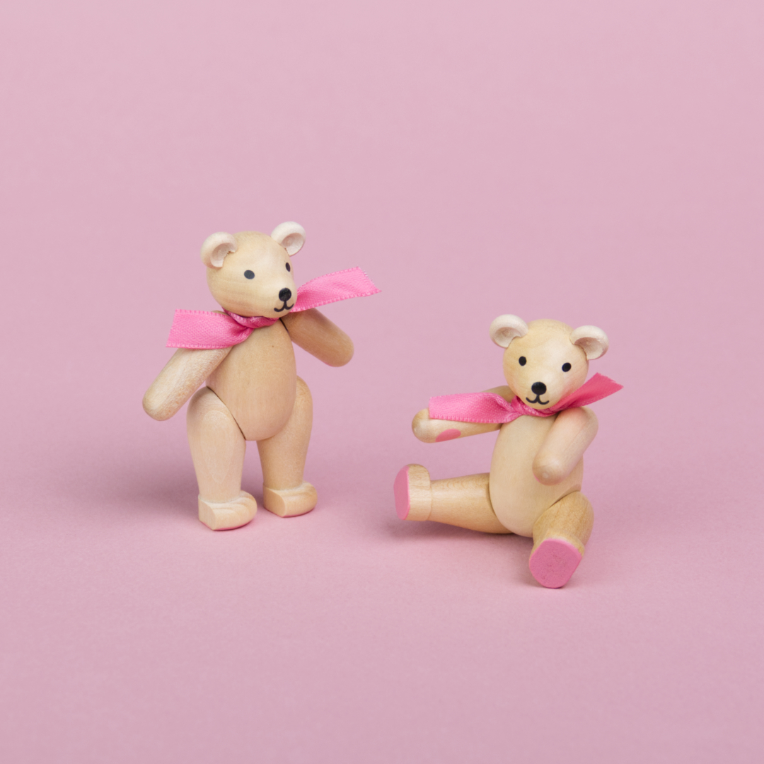 Zwei kleine Teddybären aus hellem Holz mit rosa Schleifen um den Hals, Beine und Arme sind beweglich , einer ist in einer stehenden Position zu sehen der andere sitzt und zeigt seine rosa Pfoten, beide hat eine hellere Schnauze und schwarze Augen und Nasen