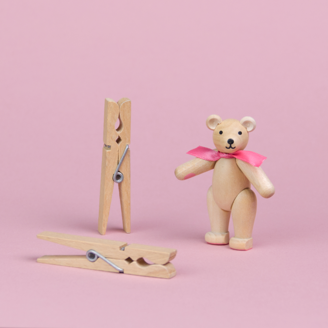 ein kleiner Teddybär aus hellem Holz mit rosa Schleife um den Hals steht neben 2 Wäscheklammern, die beiden Objekte sind annähernd gleich groß, seine Augen und Nase sind schwarz und sein Mund lächelt