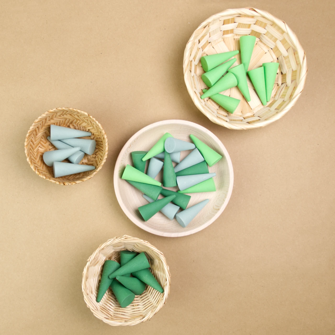 Zu sehen sind vier Behältnisse in denen sich pylonenförmige Mandalafiguren von Grapat aus Holz befinden. In der Mitte ist eine größere Holzschale zu sehen, in der sich hellgrüne, dunkelgrüne und hellblaue Figuren befinden. Rechts oben in der Ecke ist ein größeres geflochtenes Körbchen mit hellgrünen Figuren zu sehen. Links unten befindet sich ein kleines helleres Körbchen mit dunkelgrünen Figuren und links in der Mitte befindet sich ein dunkleres, kleines Körbchen mit hellblauen Figuren darin.