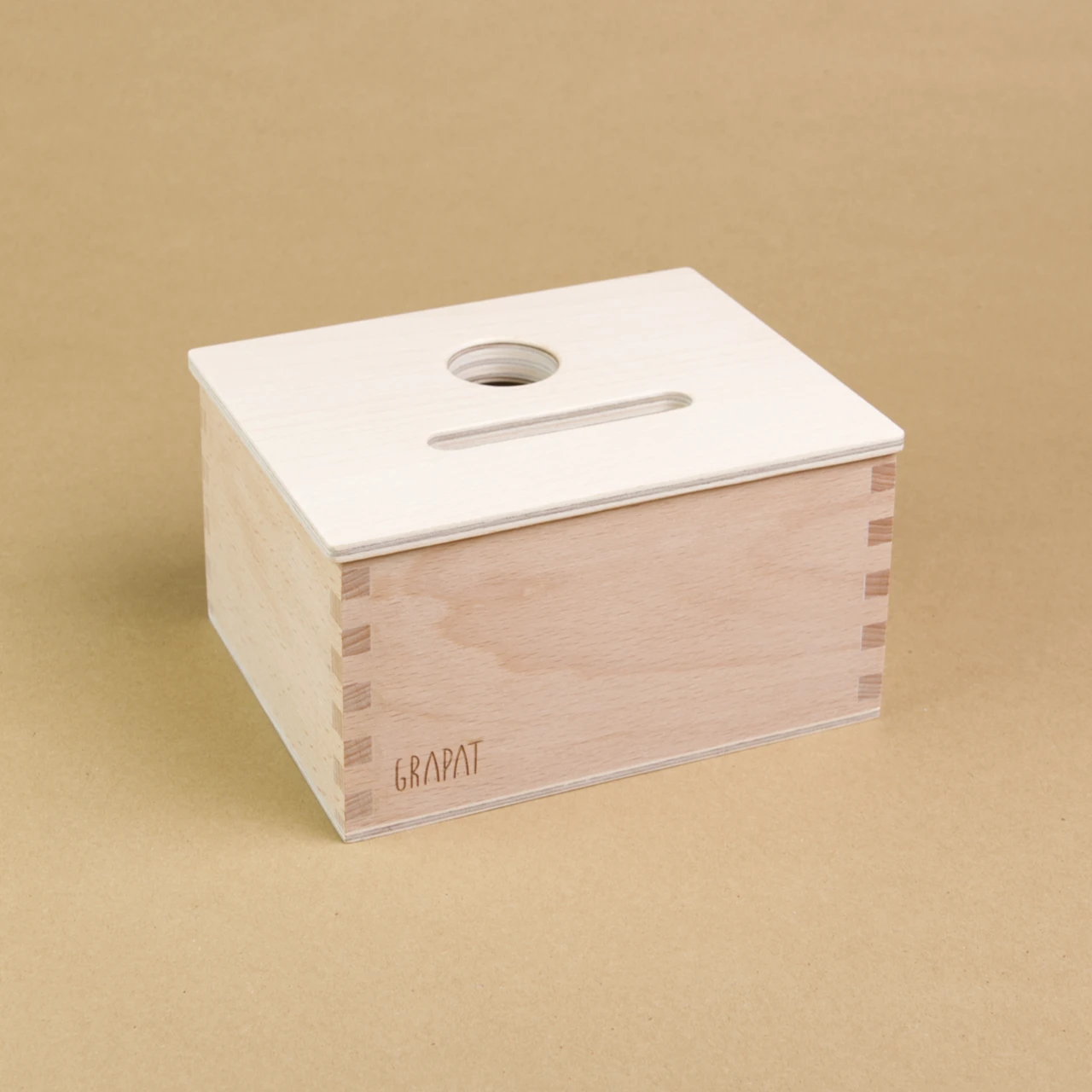 Eine rechteckige Holzbox von Grapat steht seitlich eingedreht in der Mitte, sodass drei Seiten der Box zu sehen sind. Sie hat einen Deckel in dem eine längliche und eine Runde Einkerbung sind, sodass Dinge in dieser Form in die Box geworfen werden können.