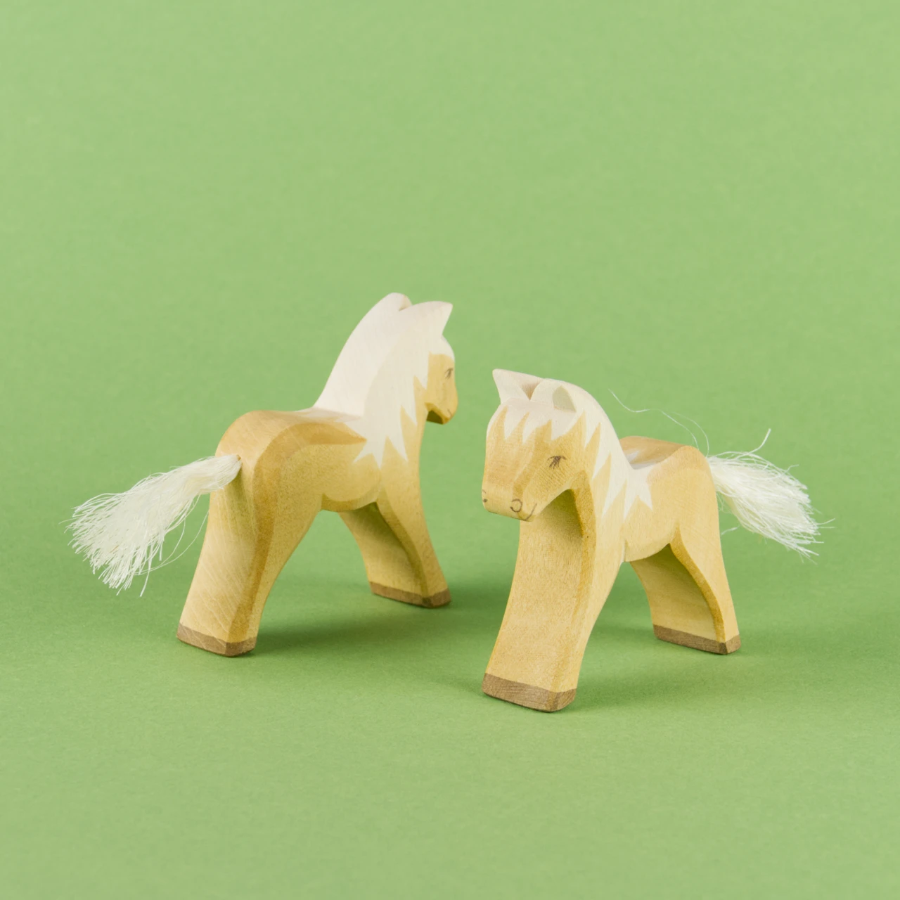 Zwei Haflinger Fohlen von Ostheimer aus Naturholz gefertigt, haben weiße Mähne und einen weißen Schweif aus Naturhaar, braune Hufen sowie braune Farbdetails. Beide schauen aneinander vorbei. Das rechts nach vorne links und das Links nach hinten rechts.