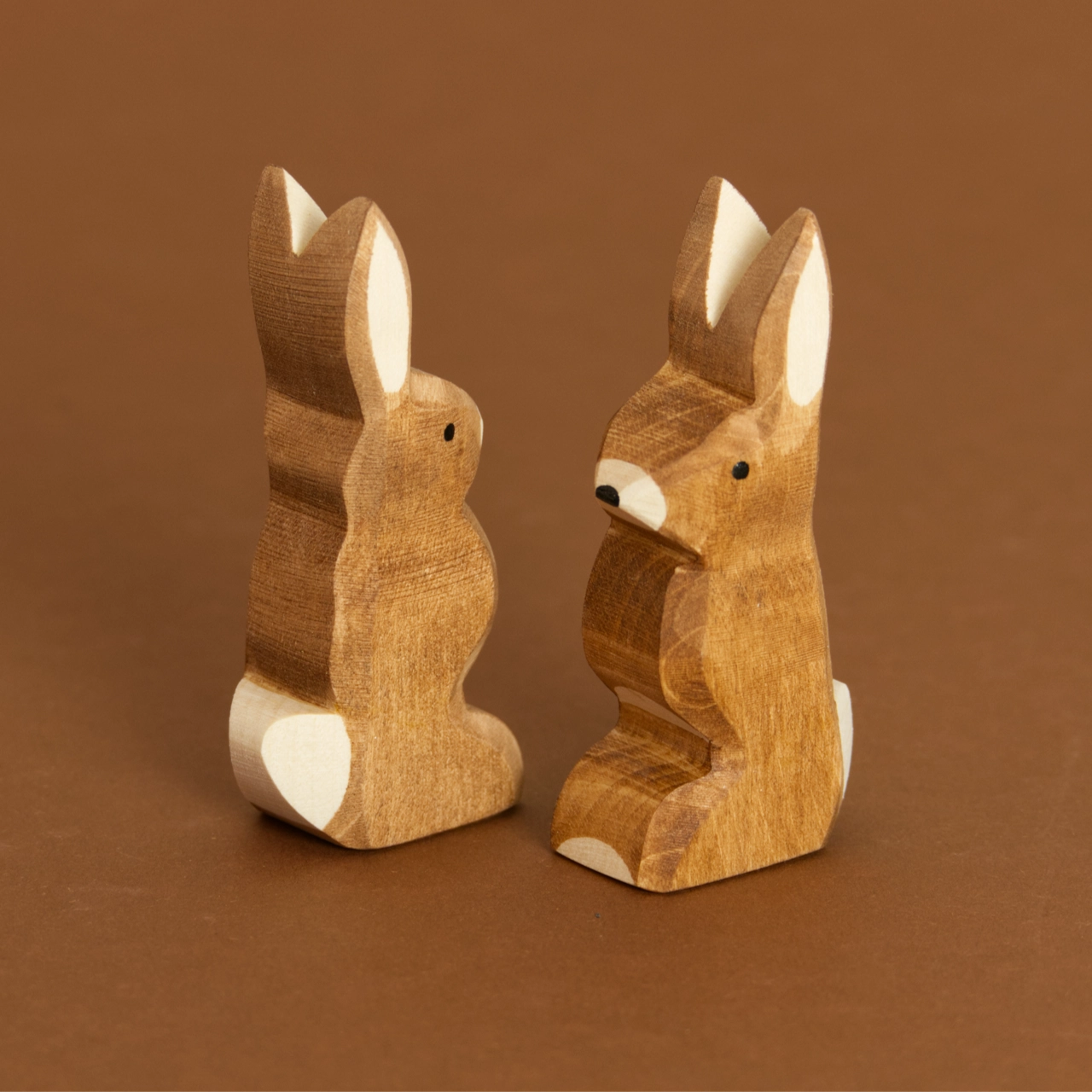 Zwei braune Hasen, mit nach oben zeigenden Ohren, schauen aneinander vorbei. Der Rechte nach vorne links und der Linke nach hinten rechts. Beide haben eine weiße Blume und eine weiße Nase mit schwarzem Farbdetail. Sie sind von Ostheimer aus Naturholz gefertigt.