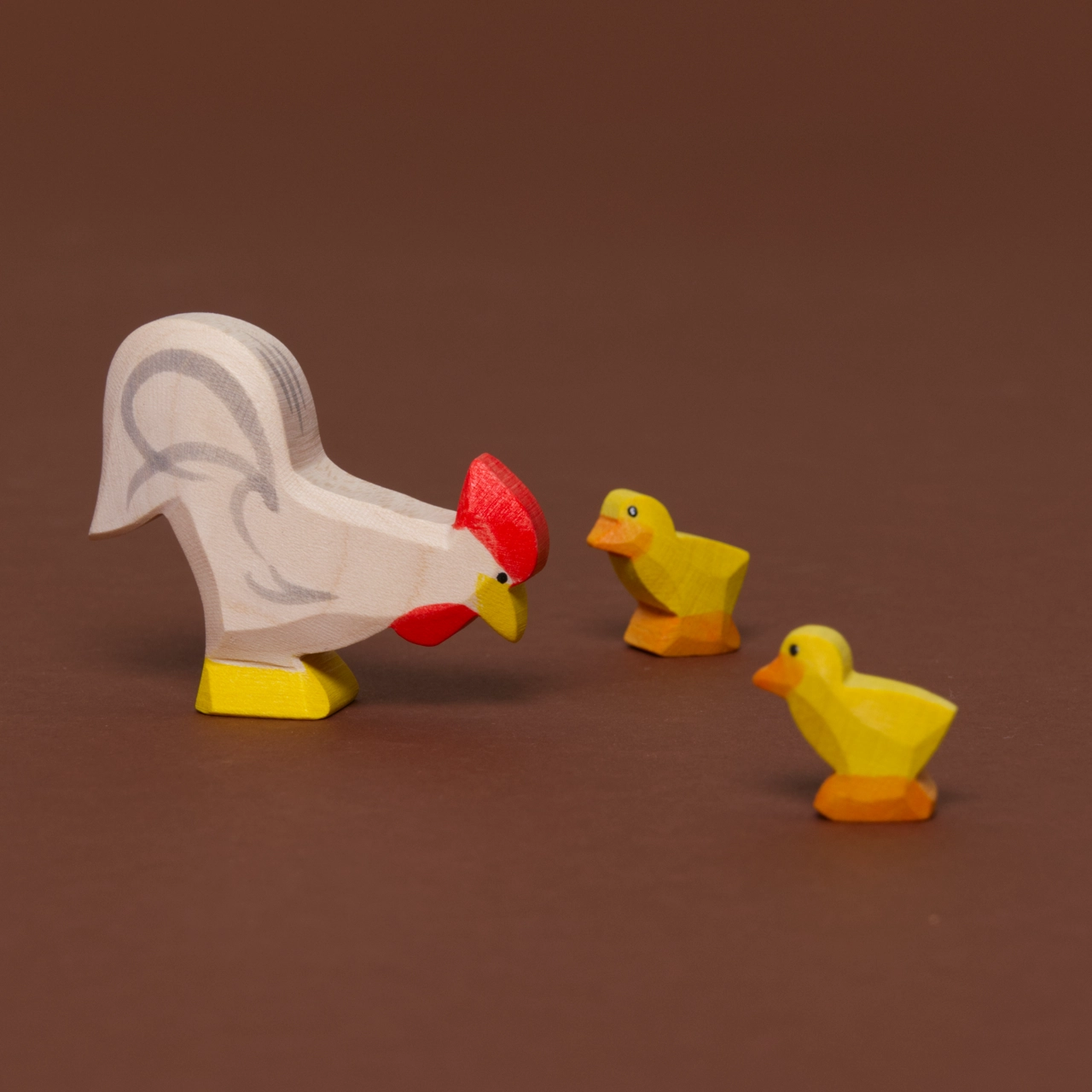 Ein weißer Hahn mit nach unten geneigtem Kopf, gelben Schnabel, sowie Füße, hat grau aufgemalte Federdetails. Es wird von zwei kleinen Küken in gelb mit orangenen Schnabel und Füßen angeschaut.