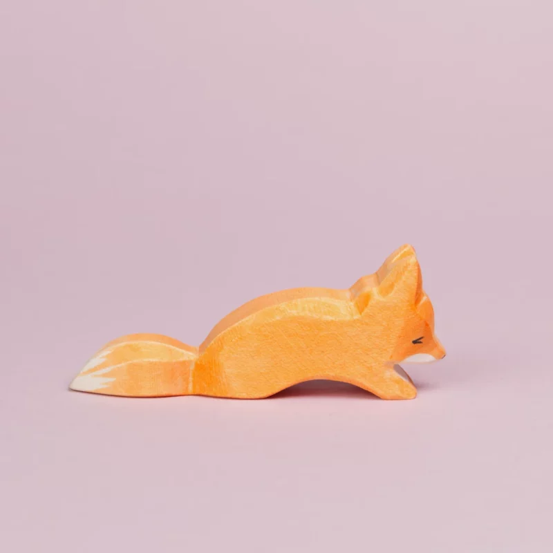 Es ist ein kleiner, schleichender Fuchs aus Holz von der Seite zu sehen, der nach rechts schaut. Sein Fell ist orangefarben und an seinem Maul und dem Ende seines Schwanzes ist es weiß. Er schaut nach unten und ist von der Marke Ostheimer.