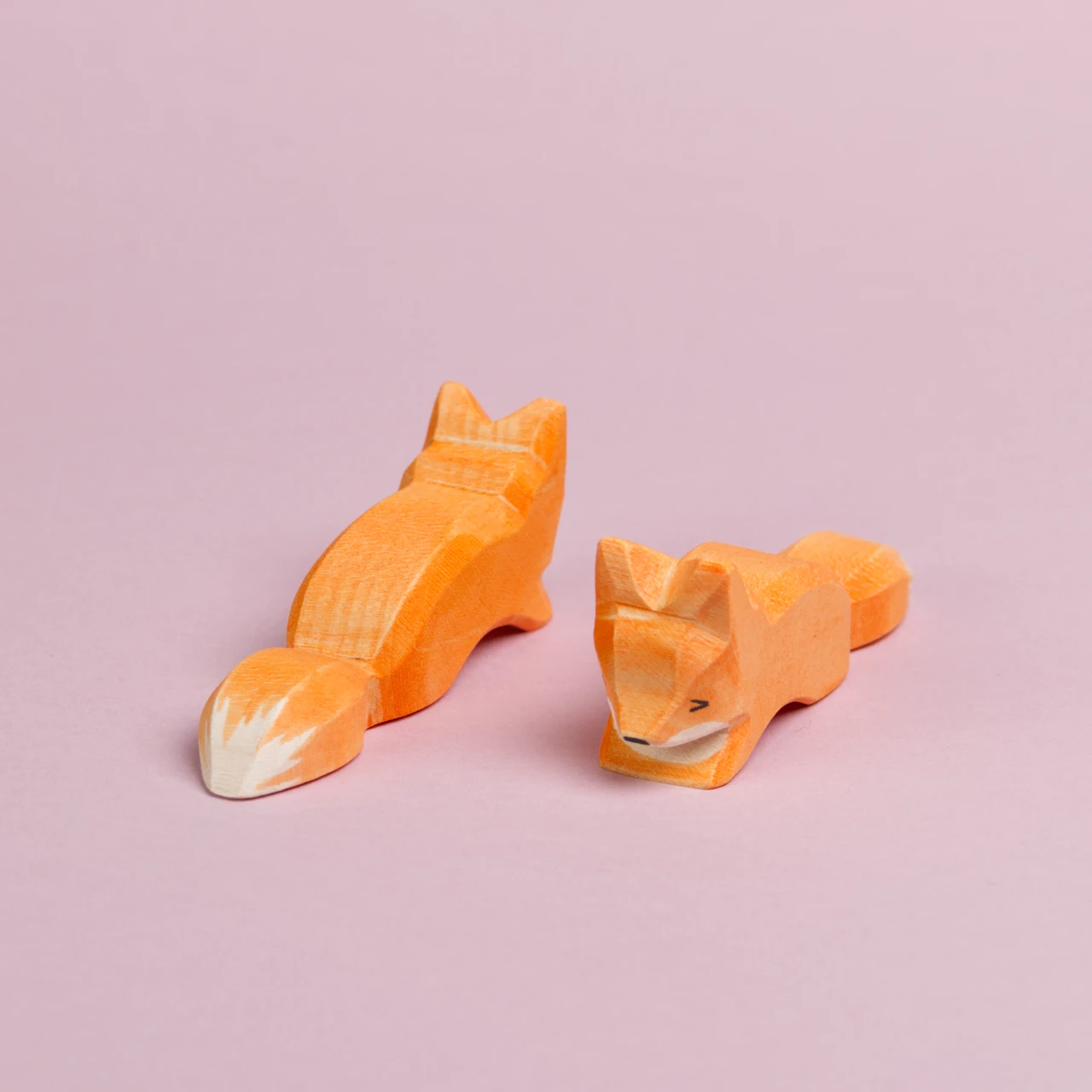 Zwei kleine, schleichende Füchse sind parallel nebeneinander zu sehen. Der rechte Fuchs schaut nach vorne links und ist mit dem Gesicht zu sehen. Der linke ist nur von hinten zu sehen und schaut nach hinten rechts.