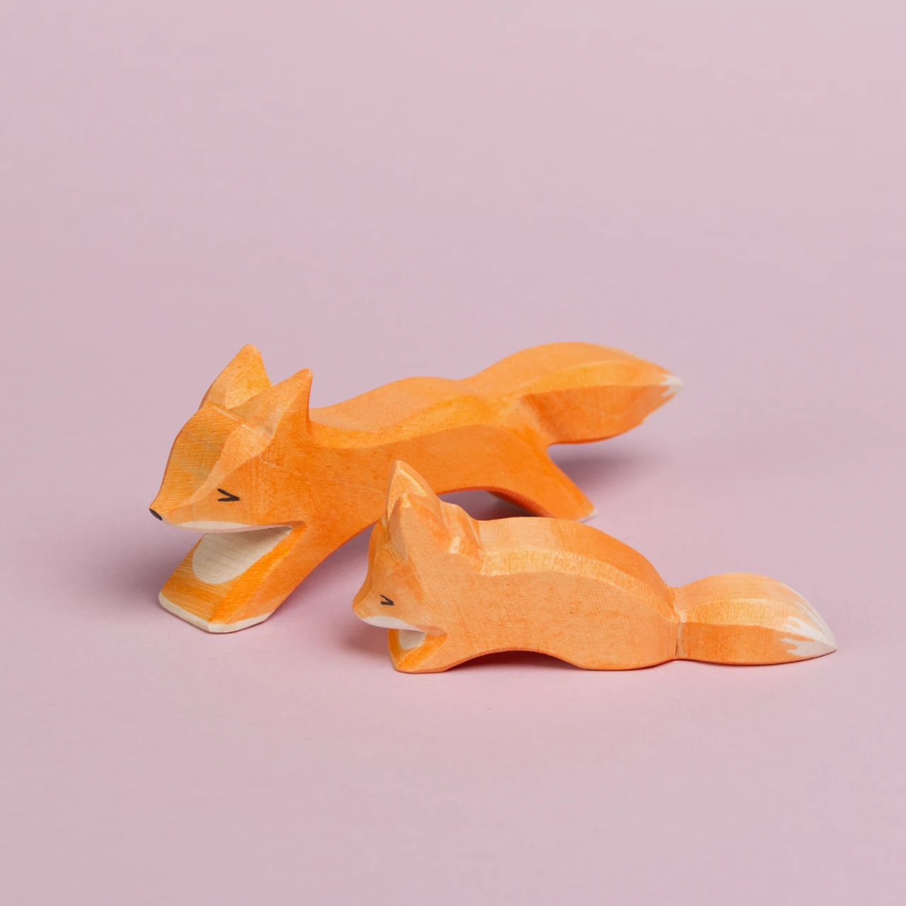 Es sind zwei Füchse aus Holz von Ostheimer nebeneinander zu sehen. Vorne ist ein kleiner schleichender Fuchs von der Seite zu sehen, der etwas weiter nach hinten versetzt ist und nach links schaut. Dahinter ist ein großer, laufender Fuchs von der Seite zu sehen, der nach vorne links schaut.