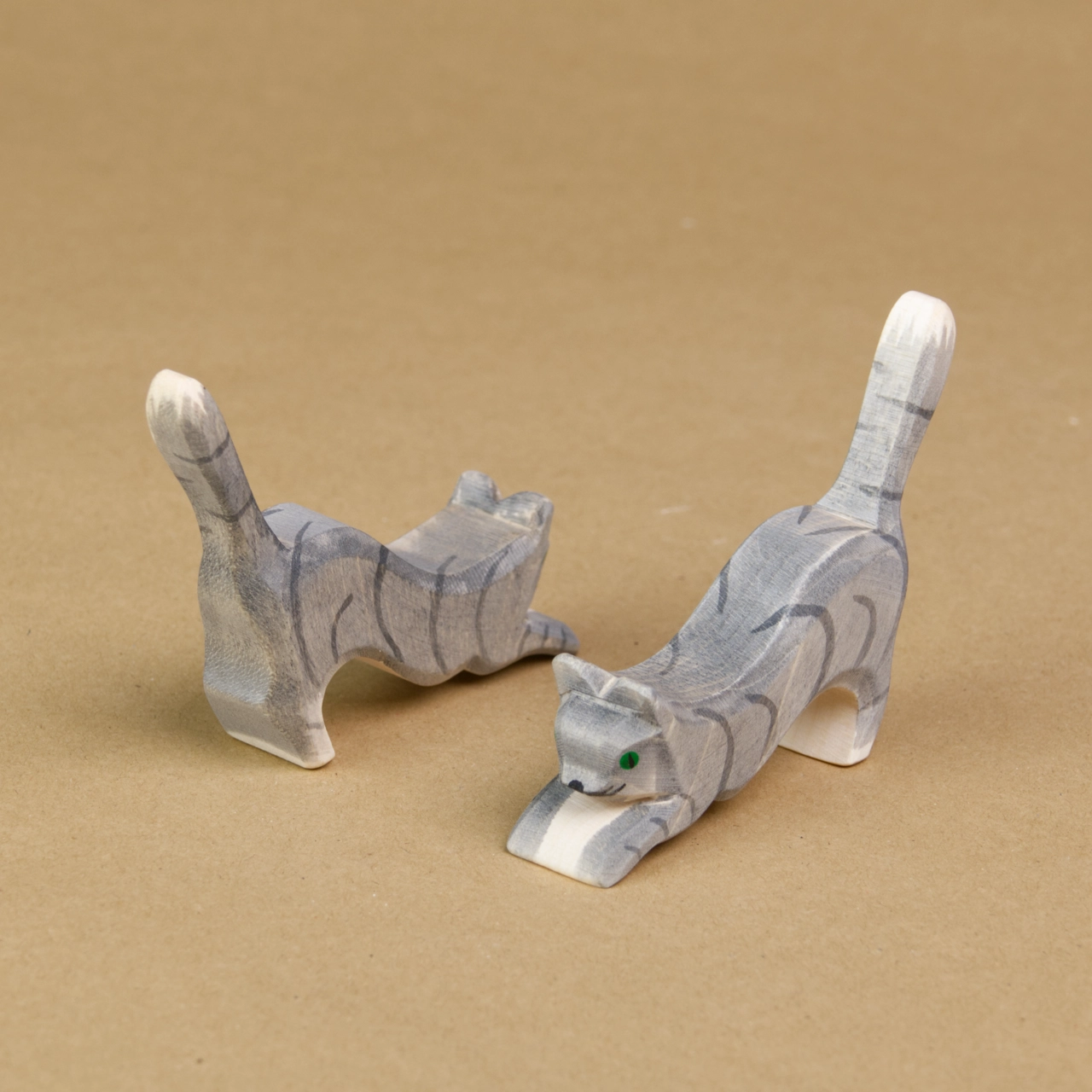 Zwei springende Katzen stehen parallel, seitlich eingedreht nebeneinander und machen einen Buckel. Die linke davon schaut nach hinten rechts und die rechte Katze schaut nach vorne links. Sie haben graues Fell und grüne Augen und sind von der Marke Ostheimer.