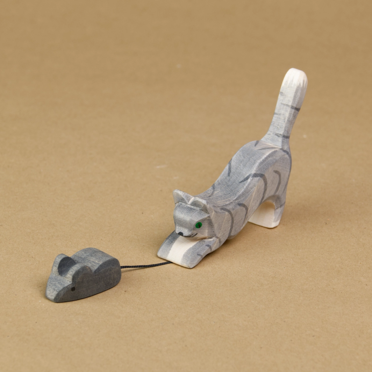 Eine springende Katze ist seitlich eingedreht zu sehen, wie sie eine Maus fängt. Sie streckt den Kopf und den Oberkörper nach unten dicht an den Boden und hält mit den Vorderpfoten den Schwanz einer Maus fest. Ihr Hinterteil streckt die Katze nach oben. Die Maus, sowie die Katze schauen nach vorne links und sind von der Marke Ostheimer.