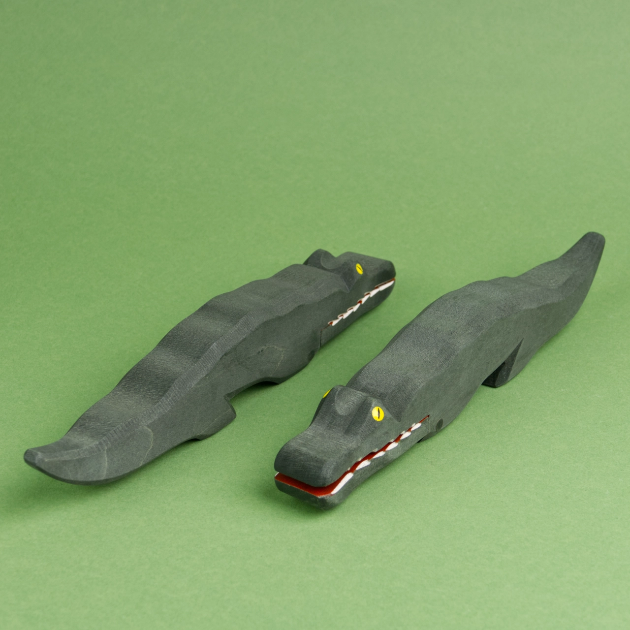 Zwei dunkelgrüne Krokodile sind schräg nebeneinander zu sehen. Das rechte schaut nach vorne links und hat das Maul leicht geöffnet, das linke Krokodil schaut nach hinten rechts und hat sein Maul geschlossen.