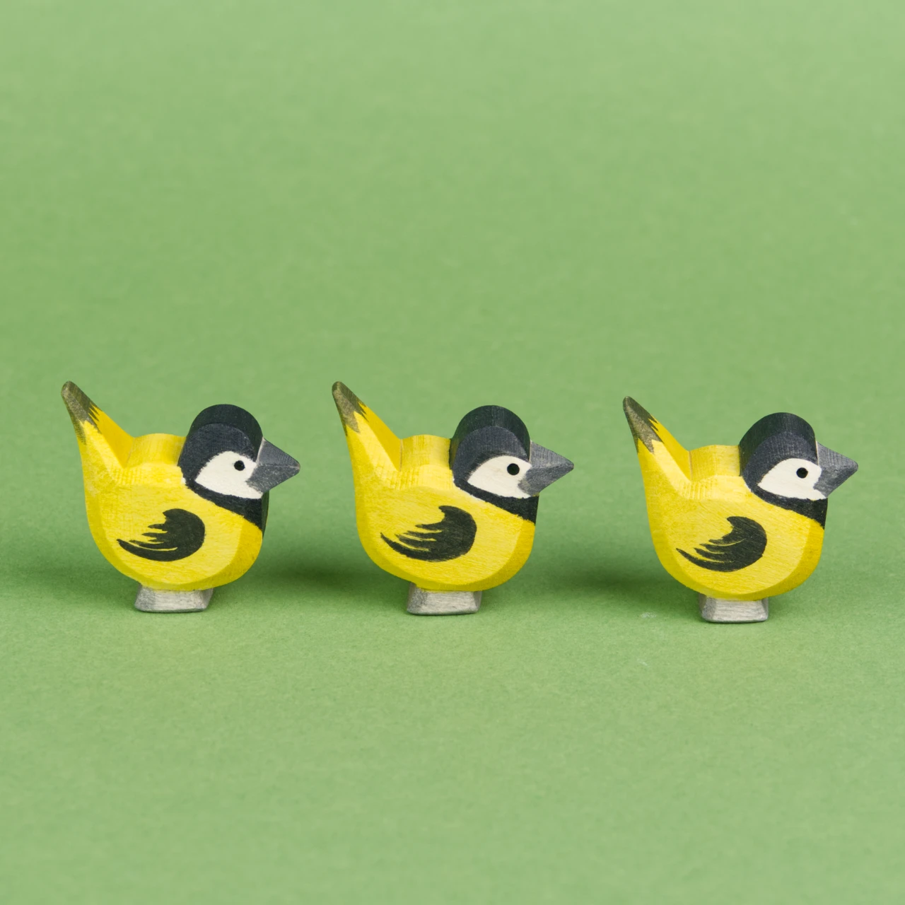 Drei Meisen aus Holz mit gelben und schwarzen Federn sind in der Seitenansicht stehend hintereinander in einer Reihe zu sehen und schauen nach rechts.