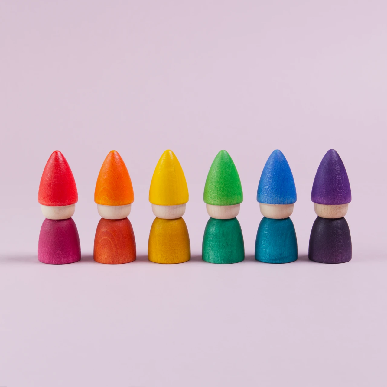 Mittig im Bild nebeneinander aufgereihte Figuren mit einer spitzen Mütze und mit farblich gleichem Oberkörper. In rot. orange, gelb, grün, blau und lila. Alle haben kein Gesicht.