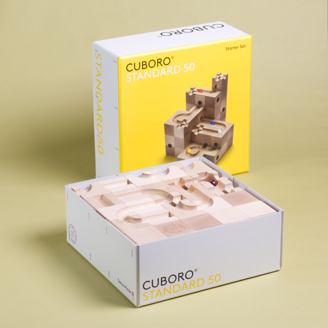 Das Standart 50 Set von Cuboro aus Buche gefertigt liegt im Produktkarton und geöffnetem Kartondeckel da, welcher im Hintergrund senkrecht aufgestellte ist.