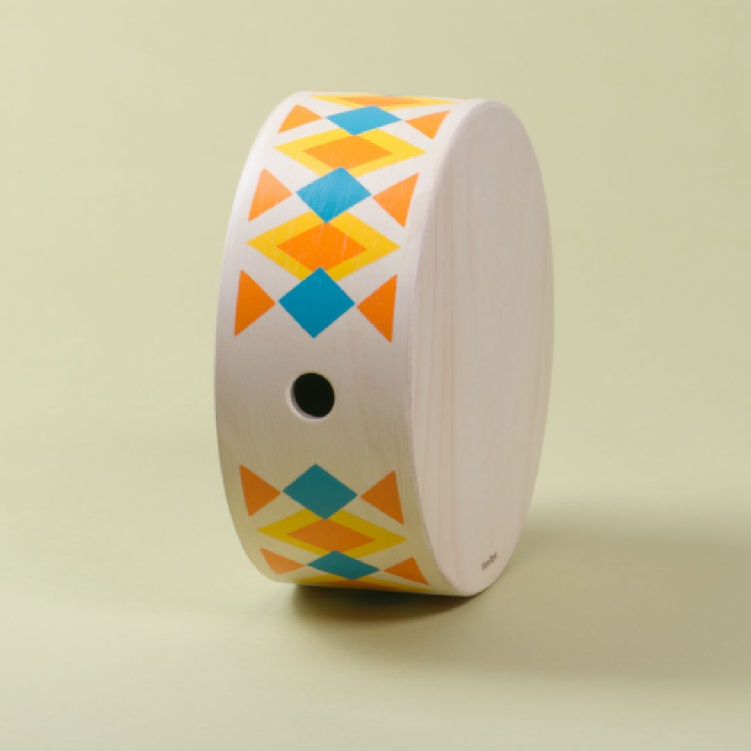 Seitenansicht mit Loch der runden Handtrommel von Plan Toys. Das helle Holz ziert ein orange, blau, gelbes Rauten bzw. Dreiecksmuster.