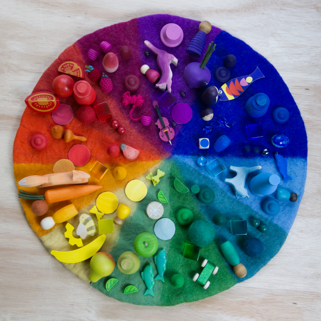 Es ist ein Farbenkreis aus Filz von der Marke Bauspiel von oben zu sehen. Die Farbfelder darauf sind nach dem Schema eines Regenbogens angeordnet. Passend zu den jeweiligen Farben, sind kleine Holzfiguren oder andere Spielwaren in der passenden Farbe auf den Feldern platziert.