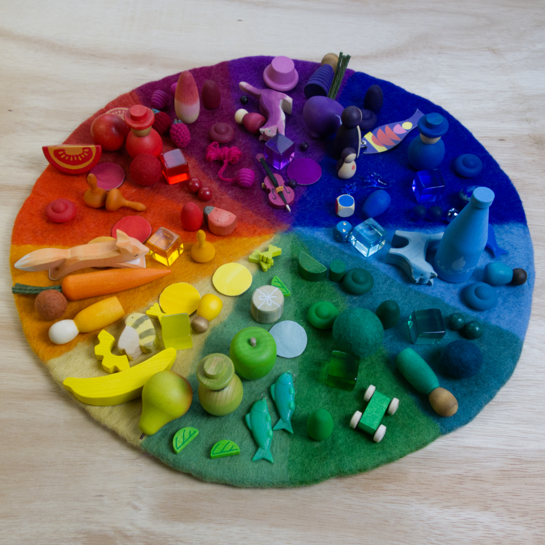 Es ist ein Farbenkreis aus Filz von der Marke Bauspiel schräg von vorne zu sehen. Die Farbfelder darauf sind nach dem Schema eines Regenbogens angeordnet. Passend zu den jeweiligen Farben, sind kleine Holzfiguren oder andere Spielwaren in der passenden Farbe auf den Feldern platziert.