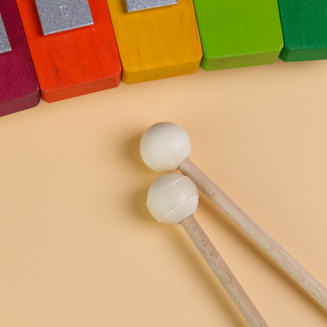 Das Regenbogenspiel von Decor-Spielzeug aus Holz enthält zwei Schlägel aus Holz, die aus einem Holzstab und einer Holzkugel darauf bestehen. Um einen der Schlägel ist ein Gummiband befestigt, sodass die Schlägel unterschiedliche Töne erzeugen.