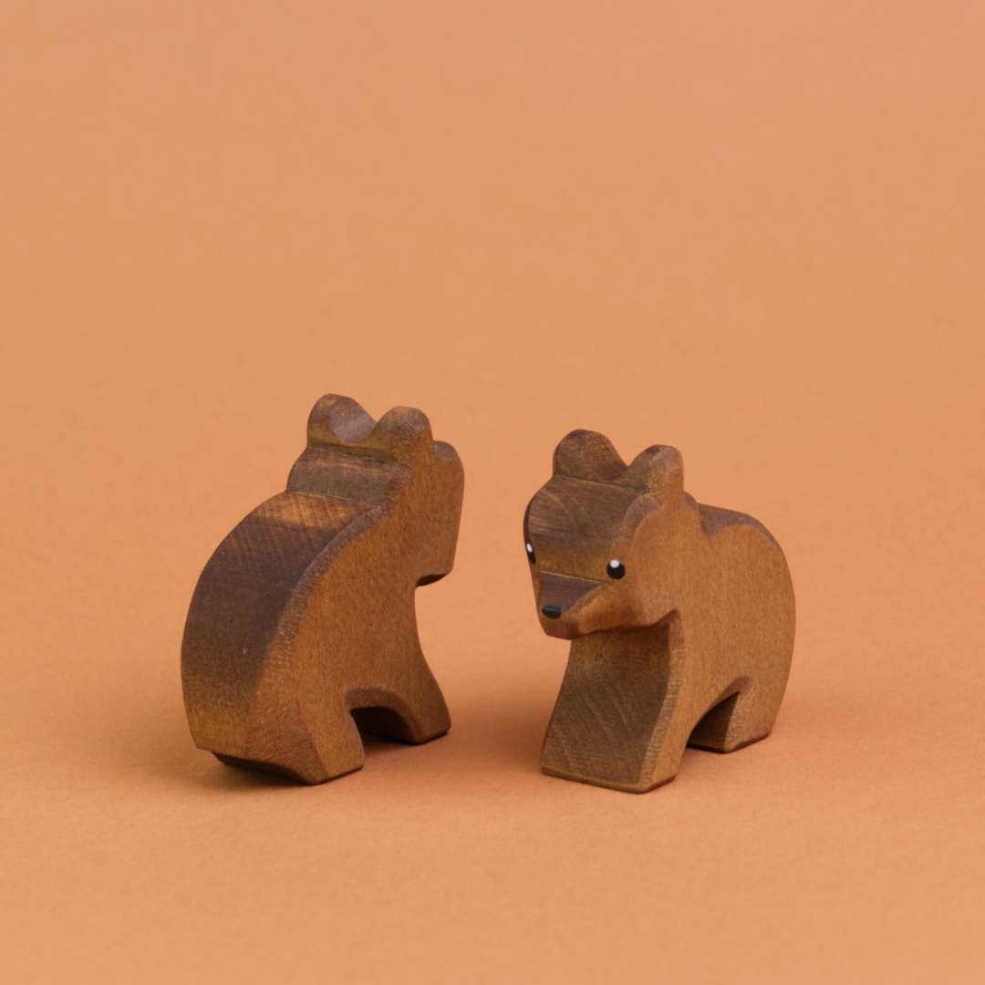 Zwei kleine Bären aus Holz sind schräg nebeneinander sitzend zu sehen. Der recht Bär schaut nach vorne links, der linke Bär schaut nach hinten rechts und ist nur von hinten zu sehen.