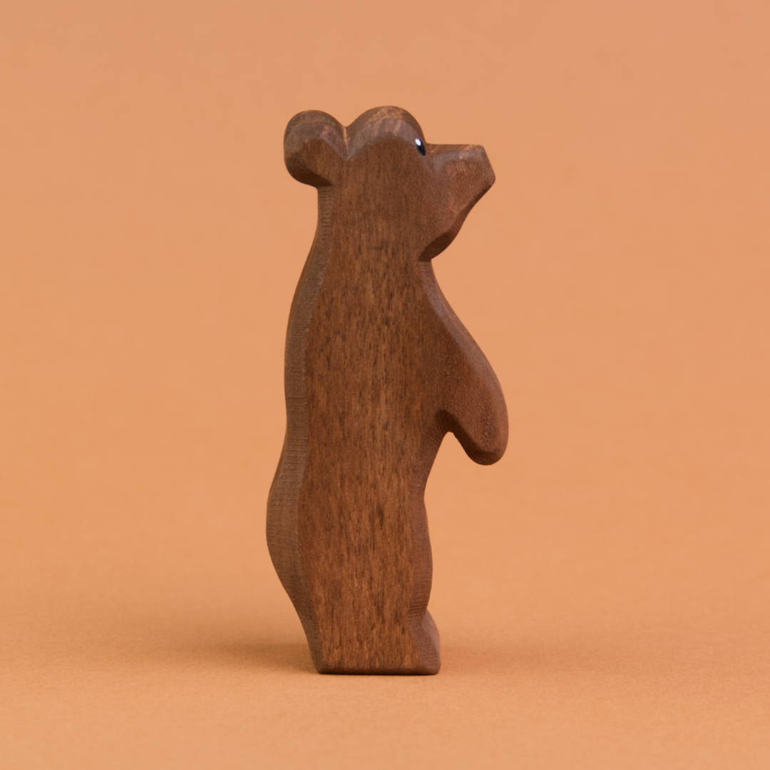 Ein kleiner Bär aus Holz ist stehend in der Seitenansicht zu sehen und schaut nach rechts, oben. Er hat dunkelbraunes Fell und große, runde Ohren. Er ist von der Marke Ostheimer.