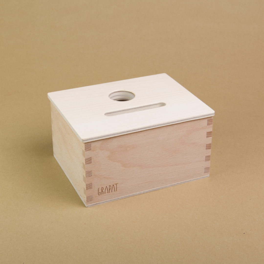 Eine rechteckige Holzbox von Grapat steht seitlich eingedreht in der Mitte, sodass drei Seiten der Box zu sehen sind. Sie hat einen Deckel in dem eine längliche und eine Runde Einkerbung sind, sodass Dinge in dieser Form in die Box geworfen werden können.