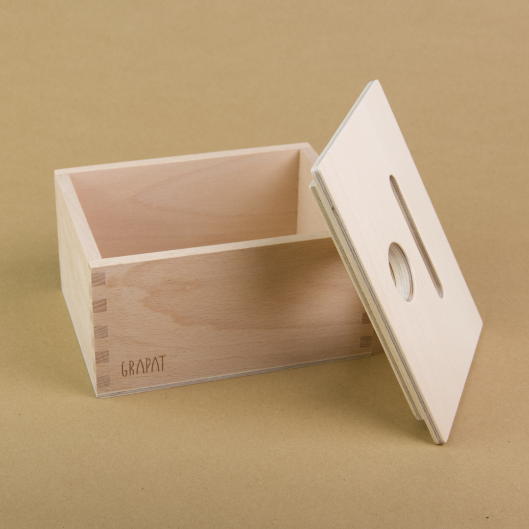 Es ist eine rechteckige Holzbox von Grapat zu sehen. Sie hat einen Deckel in dem eine längliche und eine Runde Einkerbung sind, sodass Dinge in dieser Form in die Box geworfen werden können. Der Deckel ist seitliche an die vordere, rechte Ecke der Box gelehnt.