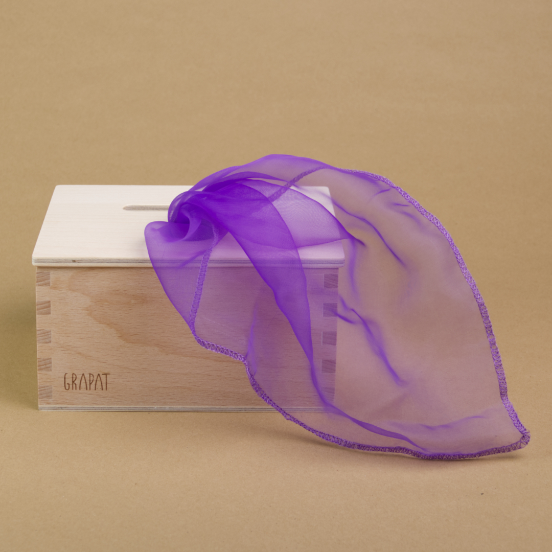 Eine rechteckige Permanente Box aus Holz steht gerade im linken Teil des Bildes. Nach rechts erstreckt sich ein violett färbendes, transparentes Tuch, das aus einer Öffnung des Deckels der Box kommt.