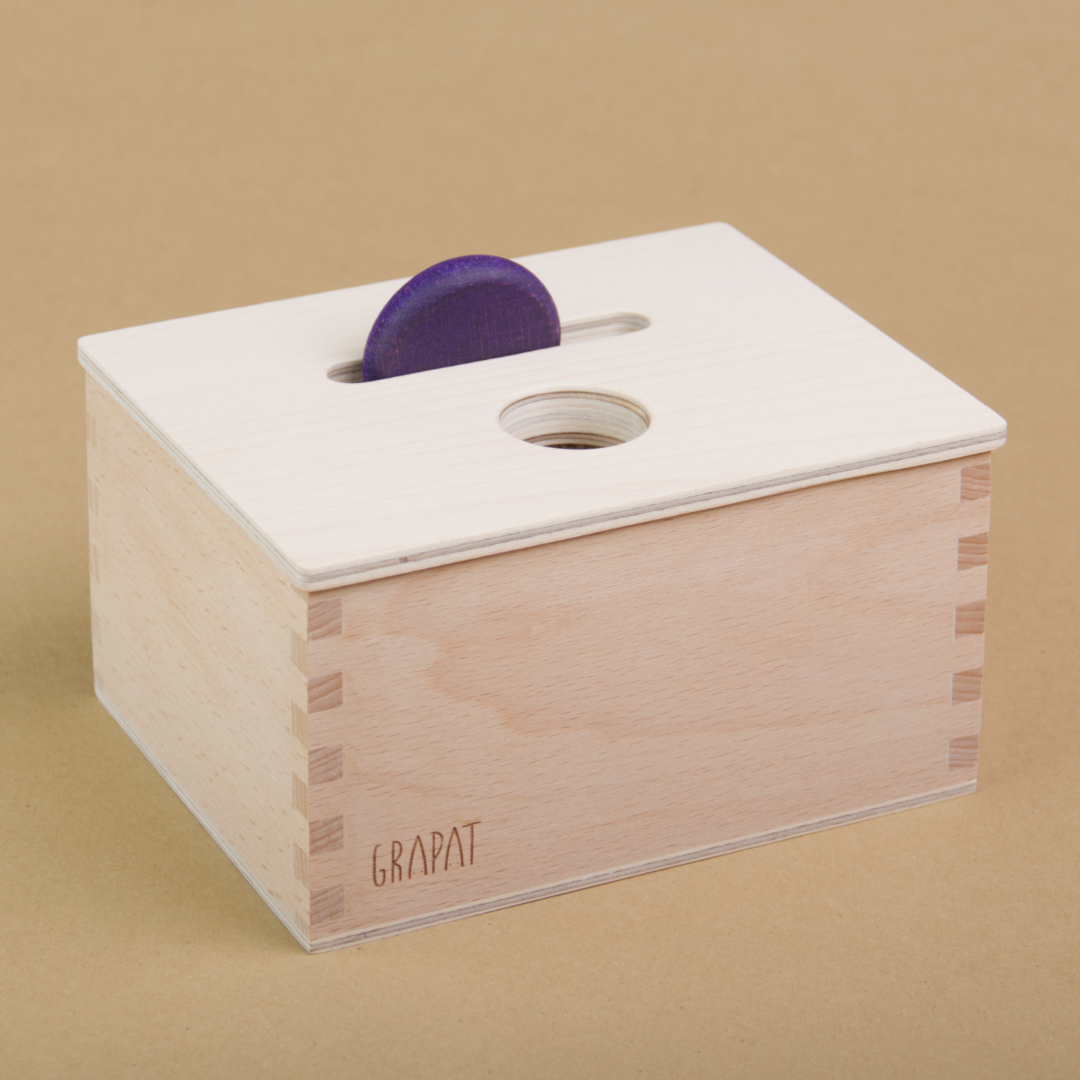 Eine rechteckige Holzbox von Grapat steht seitlich eingedreht in der Mitte, sodass drei Seiten der Box zu sehen sind. Sie hat einen Deckel in dem eine längliche und eine Runde Einkerbung sind. In der länglichen Einkerbung steckt eine violette Holzmünze.