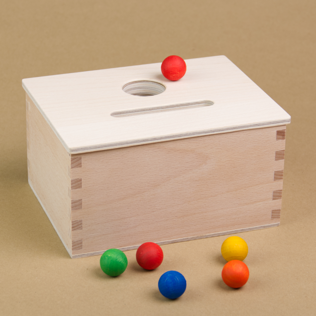 Eine rechteckige Permanence Box von Grapat aus Holz steht seitlich eingedreht da, sodass drei Seiten der Box zu sehen sind. Sie hat einen Deckel in dem eine längliche und eine Runde Einkerbung zu sehen sind. Neben der runden Einkerbung liegt eine rote Holzmurmel. Vor der Holzbox sind auch eine weitere rote, eine gelbe, orangene, grüne und blaue Holzmurmel zu sehen.