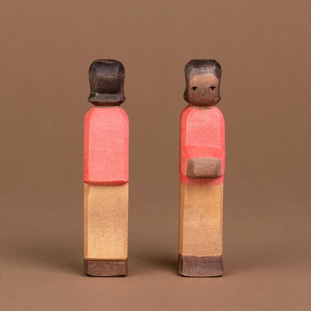 Zwei Söhne aus Holz mit dunkler Hautfarbe sind nebeneinander parallel stehend zu sehen. Der rechte Sohn schaut nach vorne und ist mit dem Gesicht zu sehen, der linke schaut nach hinten und ist nur von hinten zu sehen.