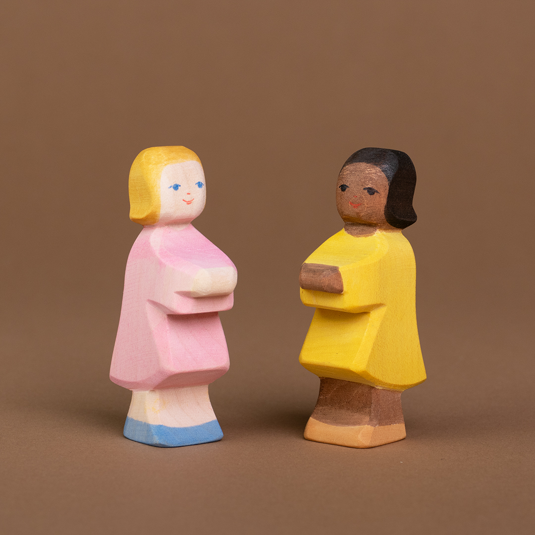 Zwei Töchter aus Holz sind nebeneinander stehend, zueinander gedreht zu sehen und schauen sich an. Die linke Tochter hat eine helle Hautfarbe und blonde Haare sowie ein rosafarbenes Kleid, die rechte Tochter hat eine dunkle Hautfarbe und dunkelbraune Haare und trägt ein gelbes Kleid. Beide sind von der Marke Ostheimer.