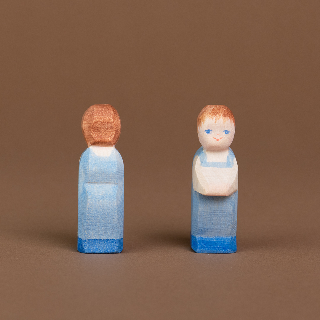Zwei Wickelkinder mit heller Hautfarbe aus Holz stehen parallel nebeneinander. Das rechte schaut nach vorne und ist mit dem Gesicht zu sehen, das linke schaut nach hinten und ist nur von hinten zu sehen.