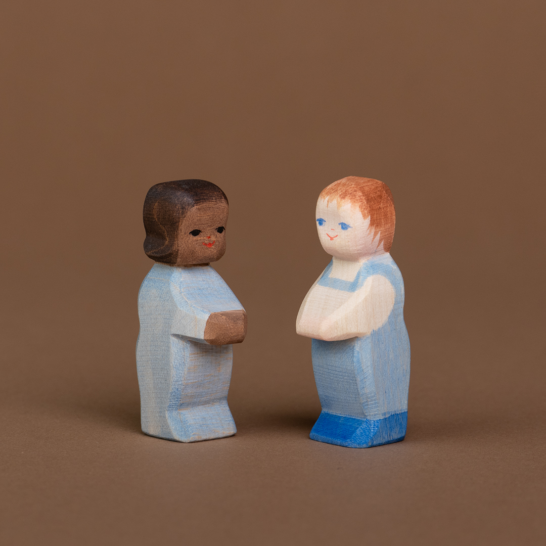 Es sind zwei Wickelkinder aus Holz zu sehen, die nebeneinander stehen und sich anschauen. Das linke hat eine dunkle Hautfarbe und trägt einen hellblauen Strampler, das rechte Wickelkind hat eine helle Hautfarbe und trägt eine hellblaue Latzhose.