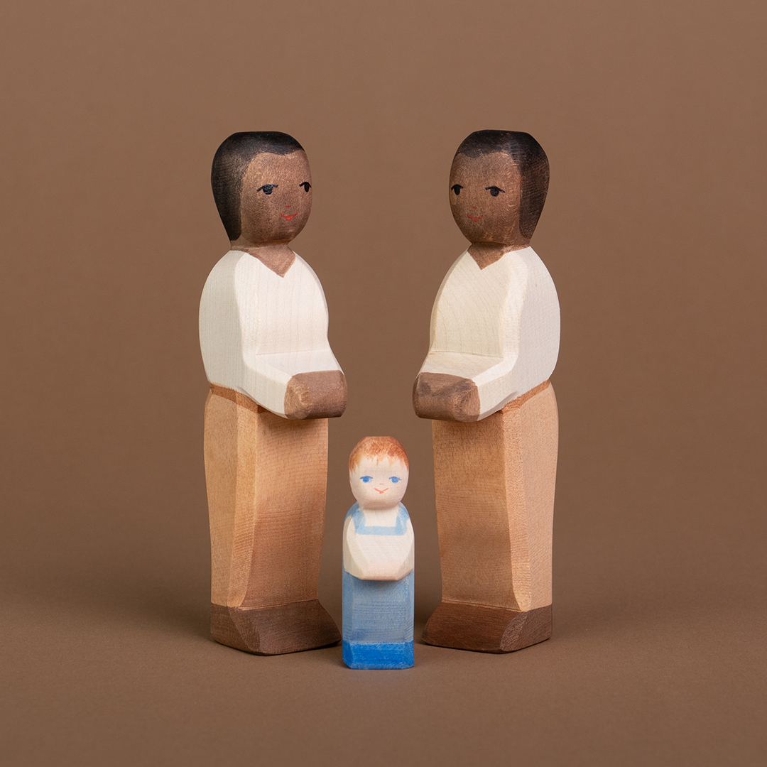 Zwei Väter aus Holz mit dunkler Hautfarbe sind einander anschauend, seitlich eingedreht zu sehen. Zwischen ihnen steht ein Wickelkind mit einer hellen Hautfarbe und schaut nach vorne.