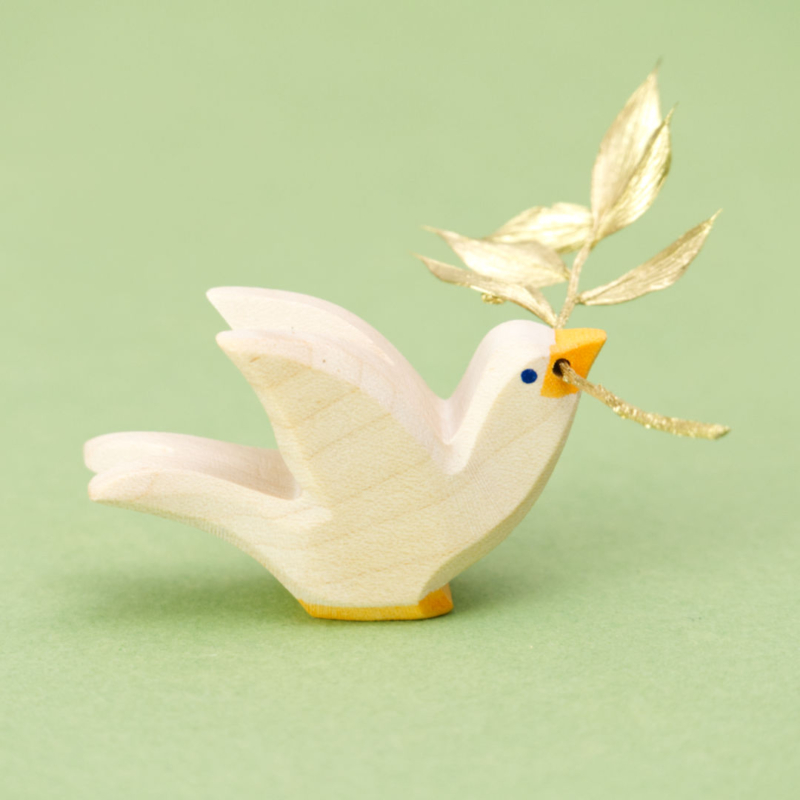 Eine hölzerne Taube der Marke Ostheimer, die nach rechts schaut. Die flach ausgesägte Figur zeigt die Silhouette eines fliegenden Vogels, die Kanten sind leicht abgerundet. Die Taube ist weiß lasiert, hat einen gelben Schnabel und gelbe Füße, die Augen bestehen aus kleinen blauen Punkten. Im Schnabel steckt ein sehr kleines Ästchen mit winzigen goldenen Blättern.