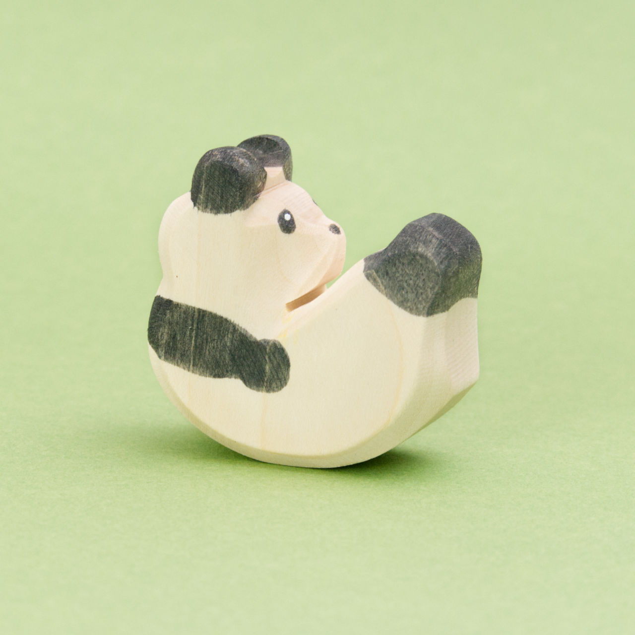 Eine kleine Panda Figur aus Holz zum Spielen von der Marke Ostheimer. Er ist von der Seite zu sehen und schaukelt auf seinem Rücken. Er hat weiß-schwarzes Fell mit der typischen Pandabären Musterung, weißer Kopf und Körper mit schwarzen Ohren, Arme und Beine, seine kleinen Augen sind schwarz umrandet.