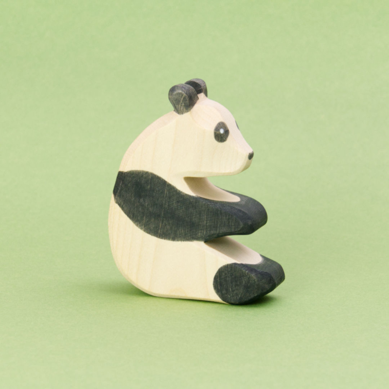 Ein sitzender Panda aus Holz zum Spielen von der Marke Ostheimer. Er ist von der Seite zu sehen und schaut nach rechts. Er hat weiß-schwarzes Fell mit der typischen Pandabären Musterung, weißer Kopf und Körper mit schwarzen Ohren, Augen, Arme und Beine. Die Holzmaserung scheint leicht durch die Bemalung und gibt der Figur einen natürlichen Charme.