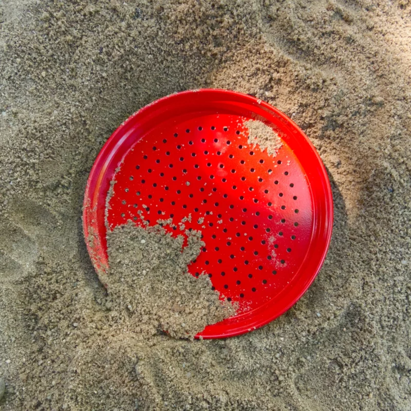 Metallsandsieb in rot von Glückskäfer. Es liegt auf dem Sand, als Draufansicht.
