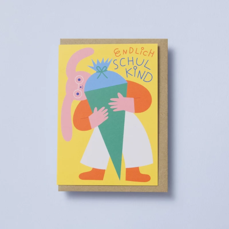 Klappkarte mit Motiv: Ein rosafarbener Hase mit runder Brille, weißer Hose, rotem Pulli und roten Socken schaut mit breitem, fröhlichem Lächeln hinter einer übergroßen Schultüte hervor. Die Schultüte ist in zarten Pastelltönen von Grün und Blau gestaltet. Im oberen Bereich der Karte steht in fröhlicher, verspielter Typografie der Schriftzug „Endlich Schulkind“. Der Hintergrund ist gelb, wodurch das Motiv freundlich und farbenfroh zur Geltung kommt – ideal für den Schulanfang. In der Klappkarte steckt ein Umschlag aus Naturpapier, von dem ein kleiner Teil sichtbar ist, da er etwas größer als die Karte selbst ist.