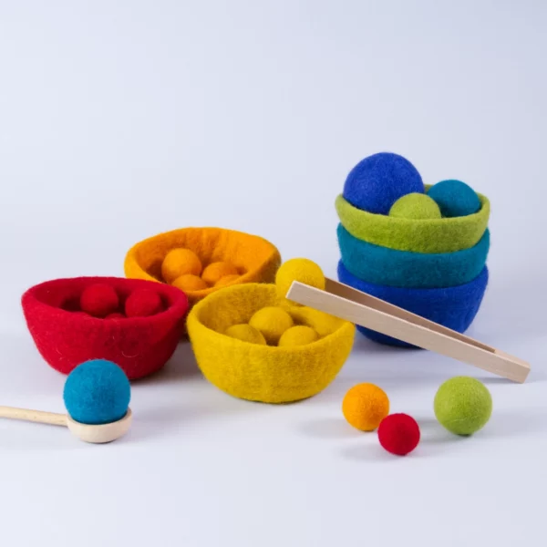 Sortierspiel Filz sorterino goki evolution mit sechs farbigen Filzschalen, farblich passenden Filzkugeln in Regenbogenfarben, Holzzange und Holzlöffel – nachhaltiges Montessori-Spielzeug zur Förderung von Feinmotorik und Farbzuordnung. Hier im Aufbau sind einige Schalen in einander gesteckt, ein Teil der Kugeln ist einsortiert. Die Holzpinzette hält einen der gelben Bälle, auf dem Löffel liegt ein Bällchen in der Farbe Türkis.